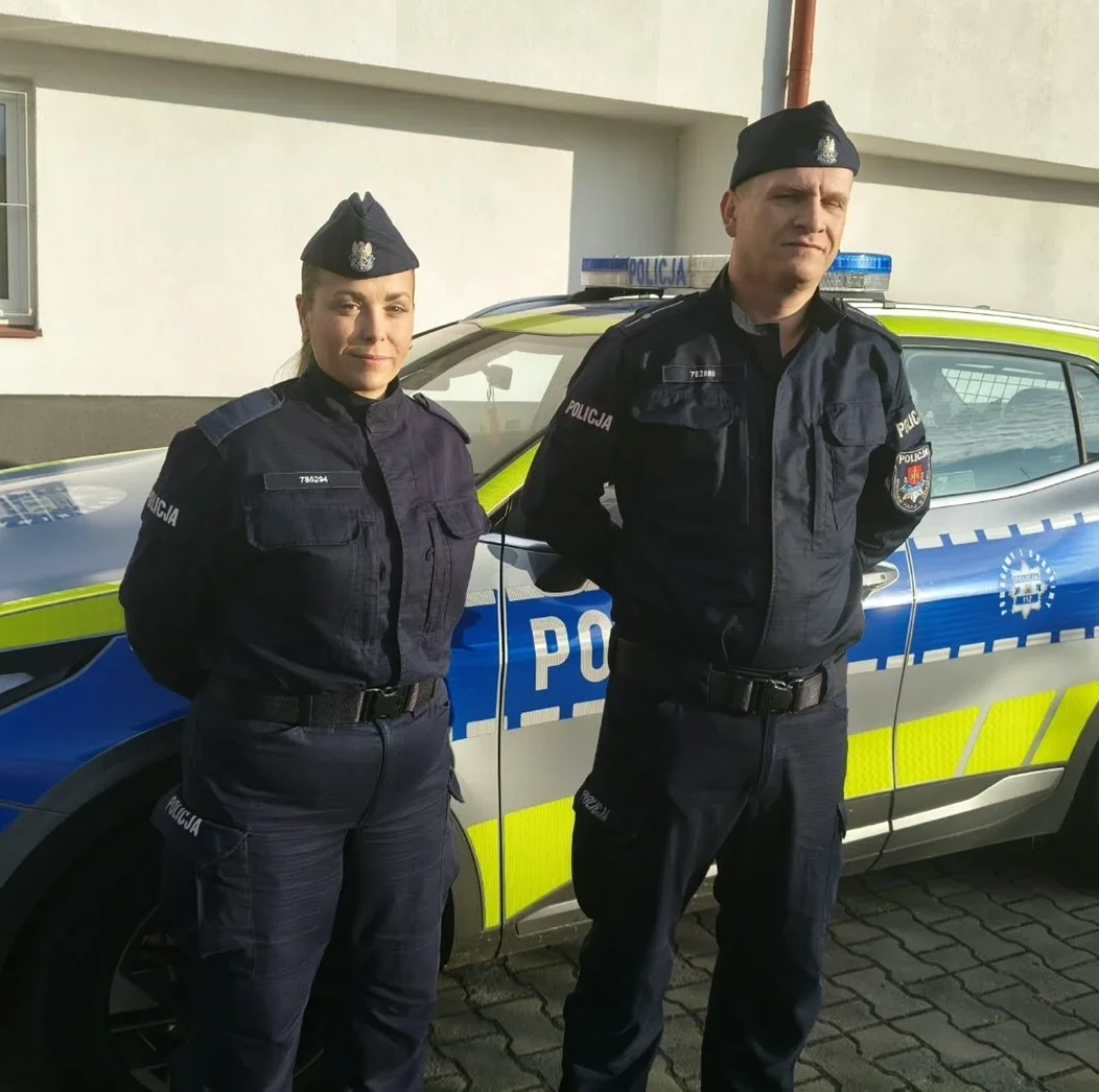 Dwoje policjantów w mundurach stoi przed policyjnym radiowozem zaparkowanym przy budynku.