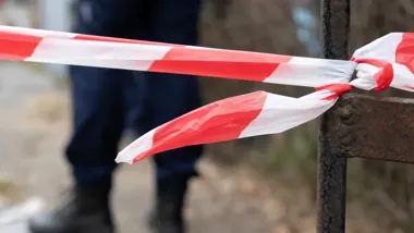 Czerwono-biała taśma policyjna przewiązana wokół rdzewiałego metalowego słupka, w tle rozmyta sylwetka funkcjonariusza w ciemnym mundurze.